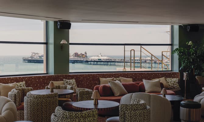 Round tables and cushioned seating face large windows overlooking a seaside pier. The interior features patterned armchairs, sofas, and small lamp fixtures, with views of the ocean and pier structures.