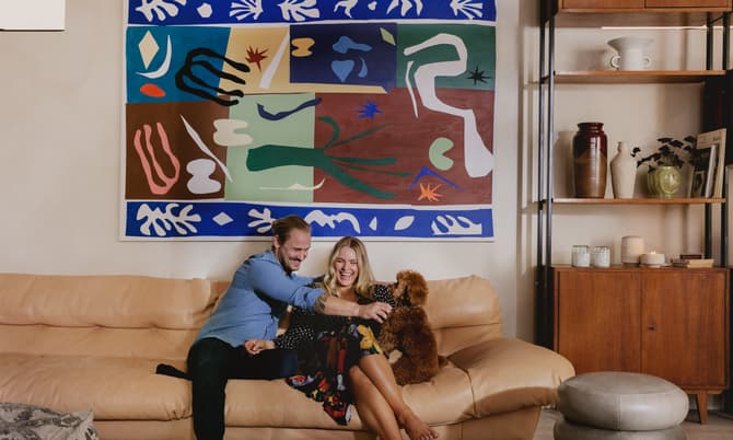 A couple on a beige sofa joyfully plays with a small brown dog in a living room adorned with a colorful abstract painting, a wooden shelf with ceramics, and candles.