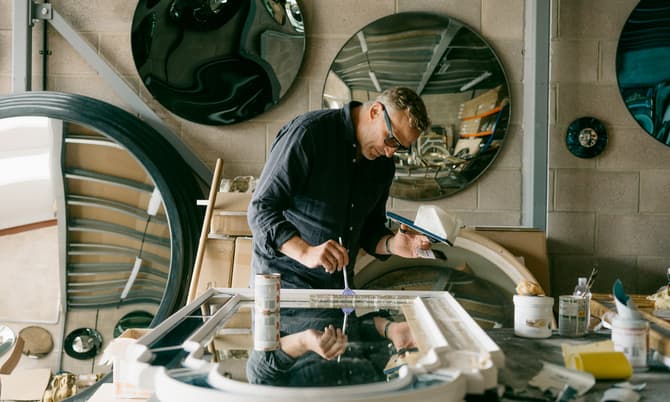 A craftsman paints a frame in a workshop with circular mirrors mounted on brick walls, surrounded by tools, boxes, and materials.