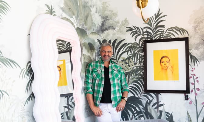 A man in a green plaid shirt and white pants stands by a wavy mirror, in a room with tropical-themed wallpaper and modern wall art.