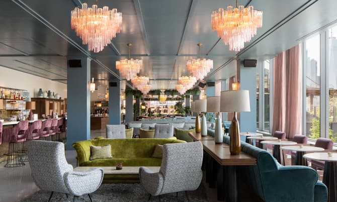 Chandeliers illuminate a modern lounge with plush seating, including a green sofa and patterned chairs, set on a carpeted floor; the space features large windows and a bar area with high stools.