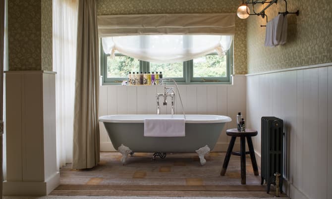 White clawfoot bathtub with a white towel draped over it, situated in a vintage bathroom. Surroundings include a wooden stool holding toiletries, a radiator, large window with a view of greenery.