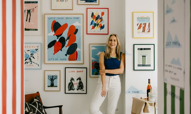 Woman leaning casually against a wall, smiling, surrounded by framed colorful artwork in a brightly lit room with red-striped wallpaper on the left.