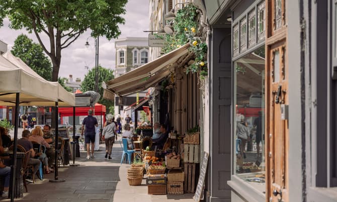 A street market featuring outdoor cafes with people seated, fruits and vegetables displayed in baskets. Passersby walk along the sidewalk. Text: "GOLBORNE ESPRESSO" on a shop sign.