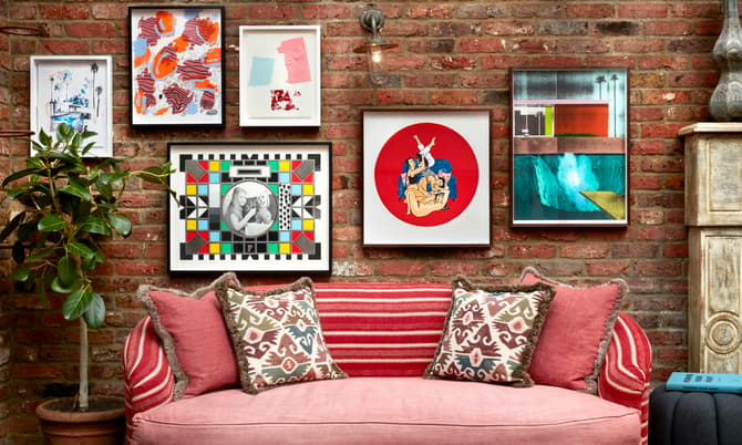 A red couch with patterned pillows sits against a brick wall displaying various framed artworks and a potted plant to the left.