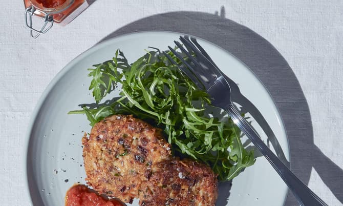 Two vegetable patties rest on a plate with arugula and red sauce, accompanied by a jar of red jam and a glass of water, on a white tablecloth. A fork is on the plate.