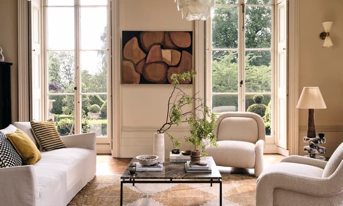 A living room with a white sofa, two armchairs, a coffee table with plants and books, surrounded by large windows overlooking a green garden, and decorated with abstract art and a chandelier.