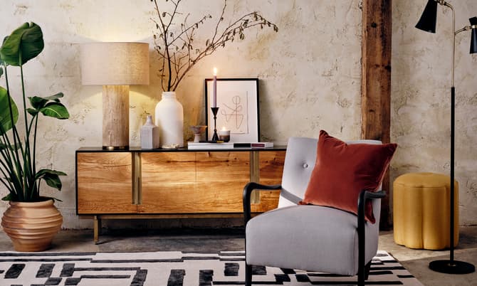 A contemporary living room features a grey armchair with a brown pillow, wooden sideboard with decor, including a lamp, plant, and candles, against a textured wall.