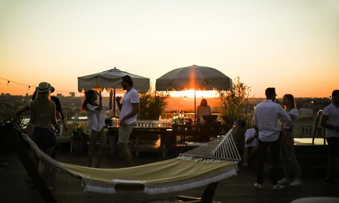 People are socializing and enjoying themselves on a rooftop at sunset, with a hammock in the foreground and umbrellas, lounge chairs, and a bar setup in the background.