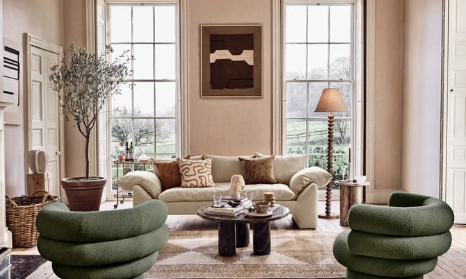 Modern living room with green, cylindrical chairs and beige sofa arranged around a central coffee table, accented by a potted tree, floor lamp, artwork, and large windows overlooking a pastoral landscape.
