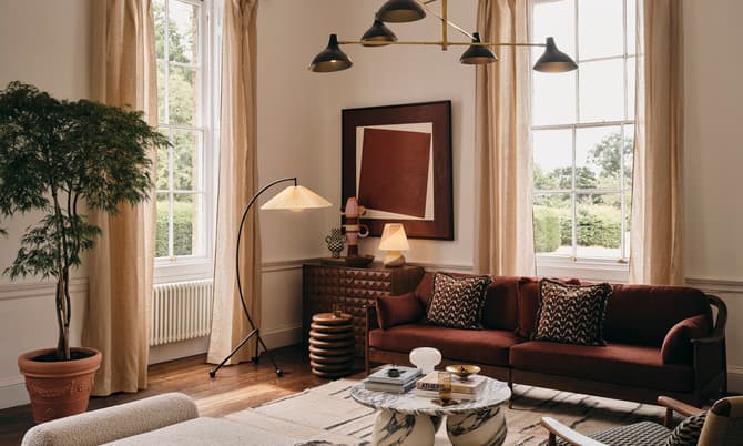 A cozy living room features a red sofa with patterned cushions, modern floor and ceiling lamps, a marble coffee table, a potted plant, and large windows with beige curtains.