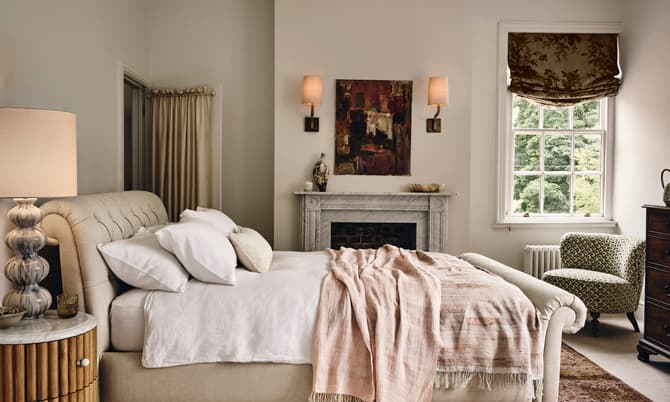 A neatly made beige bed with white pillows and a light throw blanket stands in an elegant bedroom featuring a white marble fireplace, a window with patterned drapes, an armchair, and wall sconces.