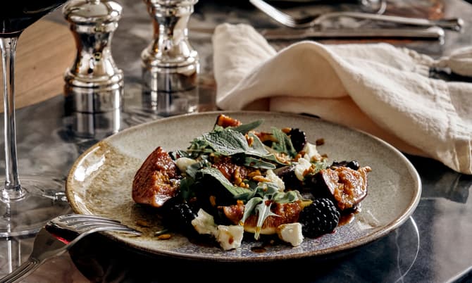 A plated salad with figs, blackberries, leafy greens, goat cheese, and nuts rests on a marble table, accompanied by a glass of red wine, cutlery, two metal shakers, and a cloth napkin.