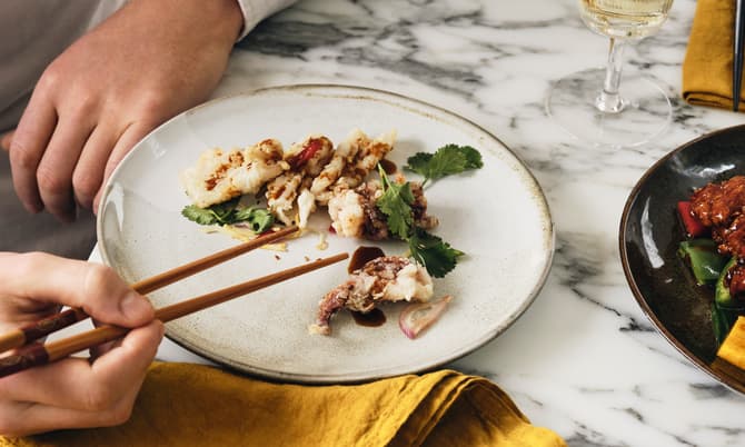 A person uses chopsticks to pick up a piece of fried food on a white plate garnished with greens, set on a marble table with a yellow napkin and a glass of white wine.