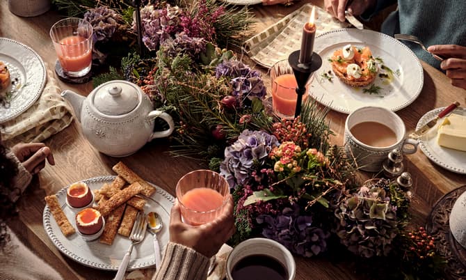 Table set for brunch with tea, juice, soft-boiled eggs, bread, and greenery centerpiece. People using utensils eating in a cozy, warmly lit environment. Plates and cups neatly arranged.