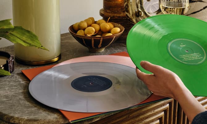 A hand holds a green vinyl record above a table. Another vinyl record lies on the table. Nearby are a candle, a fruit bowl, a plant, and a glass of white wine.
