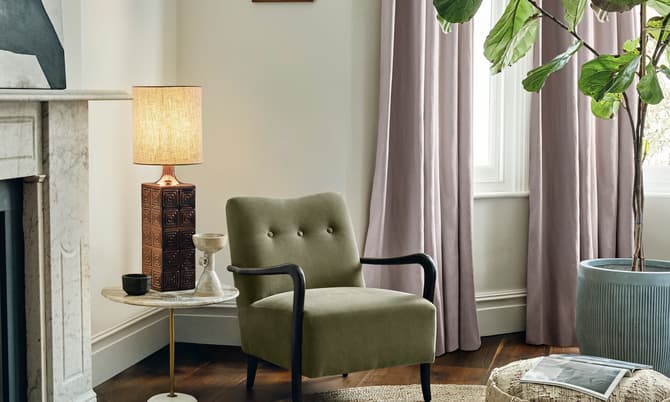 An olive-green armchair sits by a round side table with a lit lamp and decor items, placed next to a fireplace and in front of large windows with lavender curtains. There is a woven pouf with a magazine on top and a large indoor potted plant nearby.
