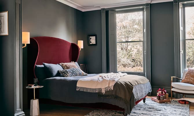 Luxurious bed with a red velvet headboard, adorned with neatly arranged pillows and a blanket, situated in an elegant, dark-walled bedroom featuring large windows with scenic views and modern décor.