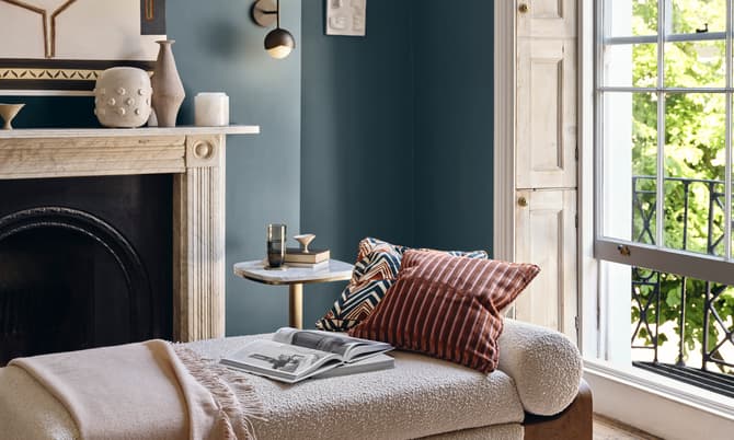 A chaise lounge with pillows and a blanket sits beside an open book and table with a glass, within a cozy room featuring a fireplace, wall art, and an open window with greenery outside.