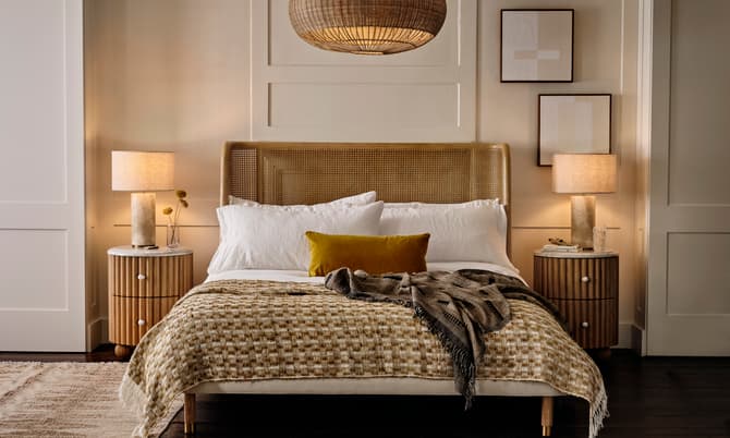A modern bedroom features a neatly made bed with white linens and a woven blanket, flanked by two wooden nightstands with lamps, framed art on the wall, and a hanging wicker light above.
