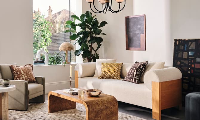 A cozy living room features a beige sofa and grey armchair with decorative pillows, centered around a wooden coffee table. Large windows, indoor plants, and art pieces create a welcoming ambiance.