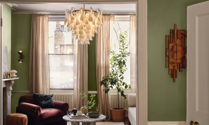 Chandelier hanging in a living room with a green armchair, coffee table, potted plants, and wall art. The space is softly lit with cream curtains and green walls.
