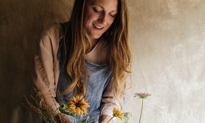 A person arranges a bouquet of flowers in a cement vase on a table, wearing an apron, with scissors nearby, against a textured beige wall.