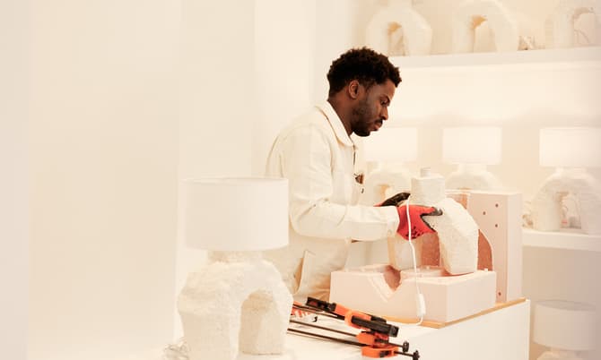 A person wearing gloves shapes a textured white lamp base on a wooden table surrounded by identical lamps and tools in a minimalist, brightly lit room.