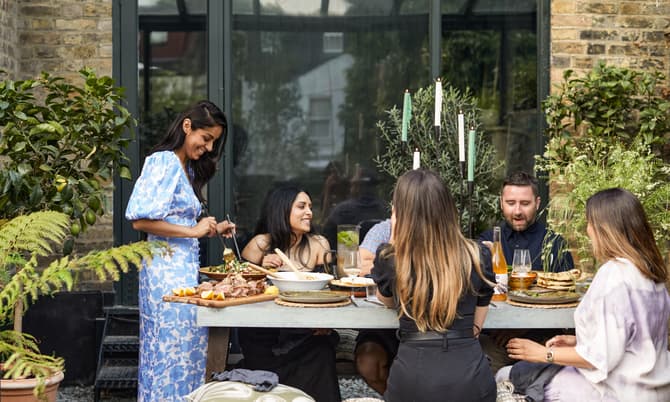 Five people sit around a table outdoors, engaged in conversation and eating. The setting is a garden with green plants and a brick wall behind them. One person serves food.