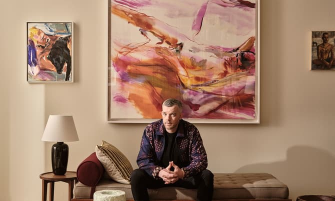 A man sits on a bench in a living room, surrounded by abstract art on beige walls, with a wooden coffee table and decor items in the foreground.