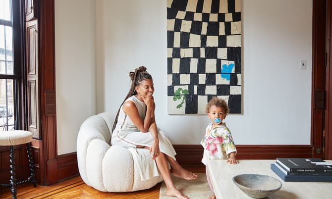 A woman sits on a cushioned chair, smiling, while a toddler with a pacifier stands nearby; they are in a cozy, light-filled room with wooden floors, near a modern art painting.