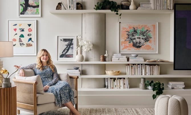 A woman in a patterned dress sits barefoot in a beige armchair in a stylish living room with art-adorned walls, bookshelves, indoor plants, and a textured rug.