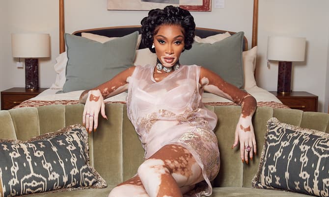 A woman lounges on a green velvet sofa in a softly lit, modern room with abstract art, wall-mounted lamps, and a marble coffee table. She's wearing a sheer, pink dress and cozy socks.