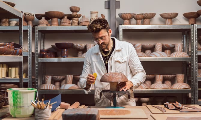 A person meticulously handcrafts a wooden bowl, using a tool for detailing, in a workshop surrounded by various shaped and patterned wooden bowls on metal shelves and a cluttered workbench.