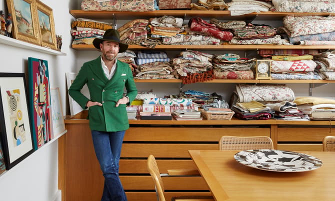 A man stands with hands in pockets, wearing a green blazer and hat, in front of shelves filled with folded fabrics and books, beside a wooden table with a decorative dish.