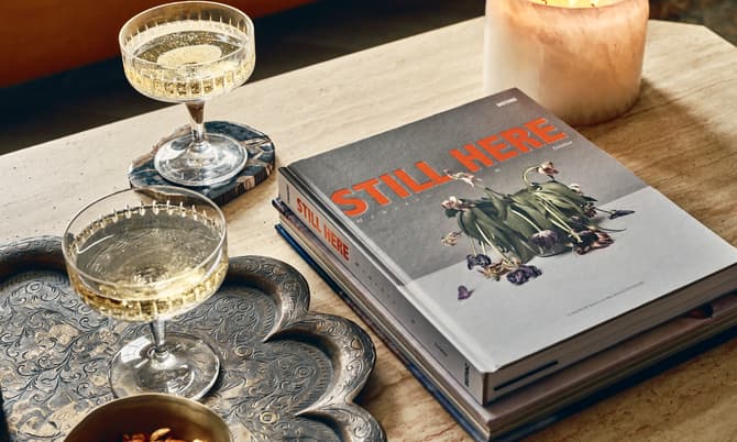 Two champagne flutes on coasters, a lit candle, a brass bowl of nuts, and books titled "STILL HERE" rest on a carved metal tray and a wooden table.