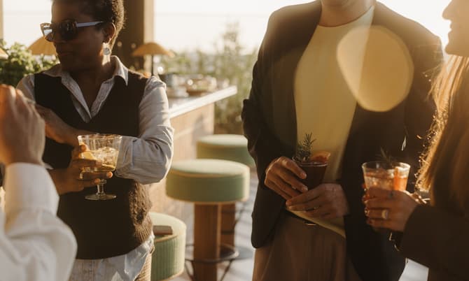 People socialize while holding cocktails, standing in a warmly lit outdoor lounge area with bar stools and plants.