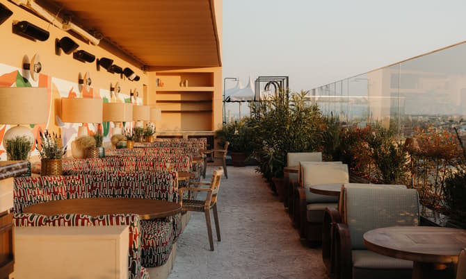 Rooftop restaurant features patterned chairs and round wooden tables set along a decorative wall with lamps and plants, offering an airy and lush ambiance with city views.