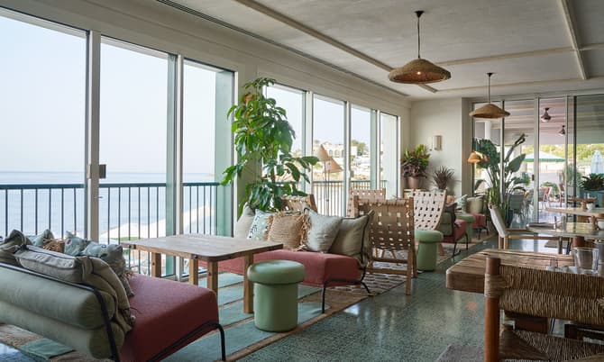 Seating area with cushions and small tables, overlooking the ocean through large windows. Indoor plants are scattered around, and pendant lights hang from the ceiling in a cozy lounge setting.