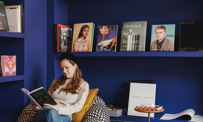 A woman reads a book while sitting in a patterned chair within a dark blue bookstore. The backdrop includes shelves with various books and a neon sign that reads "Claire de Rouen BOOKS."