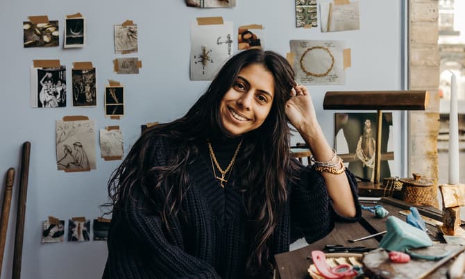 A woman with long hair and a blue sweater sits at a desk, smiling, with artwork and photos on the wall behind her in a well-lit room by a window.