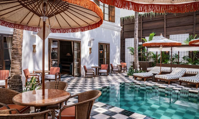 Poolside lounge chairs with umbrellas surround an empty, turquoise-tiled swimming pool. The area, bordered by tropical plants and a white building with wooden doors and windows, features wicker furniture and checkered flooring.