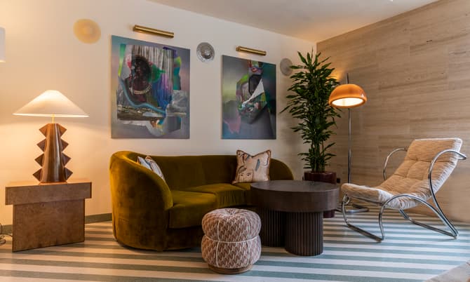 Mid-century modern living room featuring a green velvet sofa with decorative pillows, abstract wall art, a geometric table lamp, a striped ottoman, a round coffee table, a potted plant, and a unique lounge chair.