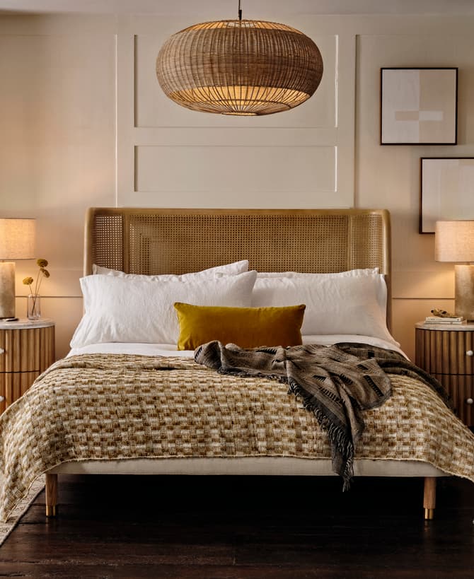 A modern bedroom features a neatly made bed with white linens and a woven blanket, flanked by two wooden nightstands with lamps, framed art on the wall, and a hanging wicker light above.