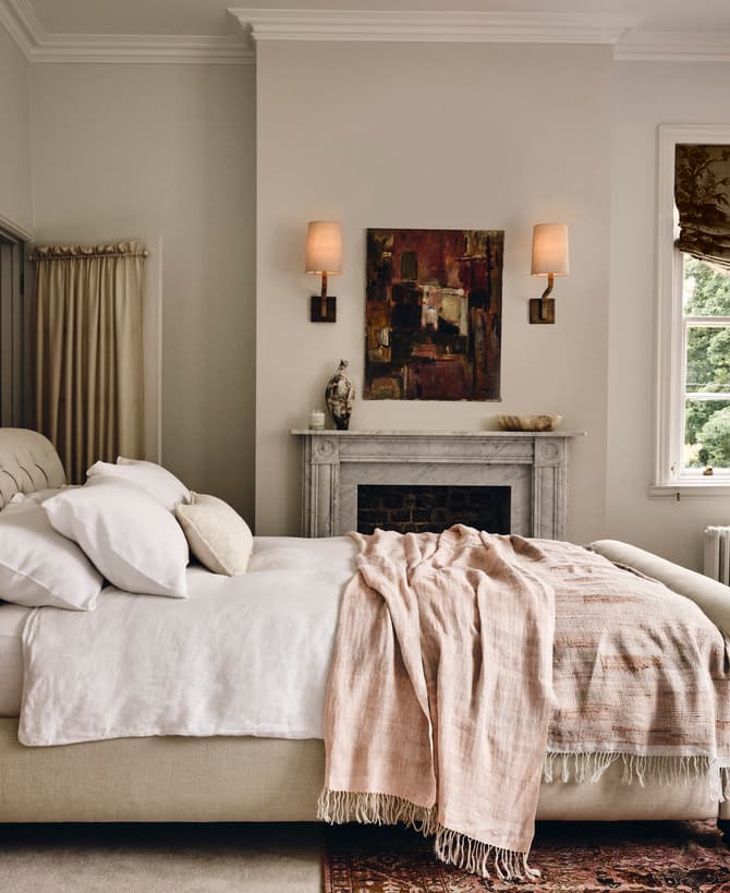 A neatly made beige bed with white pillows and a light throw blanket stands in an elegant bedroom featuring a white marble fireplace, a window with patterned drapes, an armchair, and wall sconces.