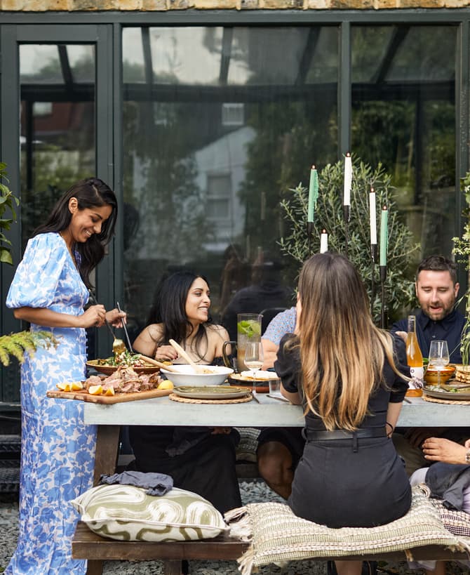 Five people sit around a table outdoors, engaged in conversation and eating. The setting is a garden with green plants and a brick wall behind them. One person serves food.