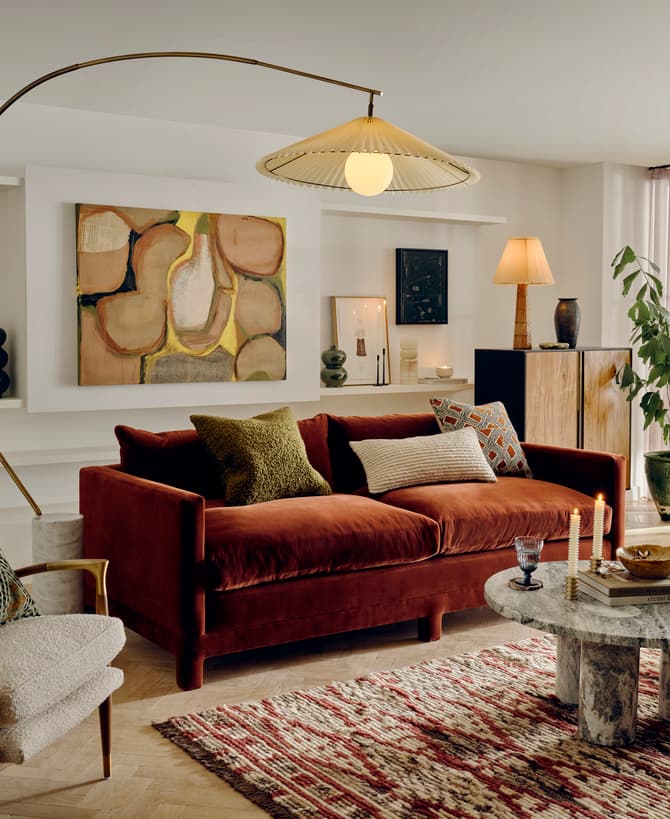 A cozy living room features a maroon velvet sofa with colorful pillows, a modern lamp, abstract art, a marble coffee table with books and candles, and a lush potted plant by sheer windows.
