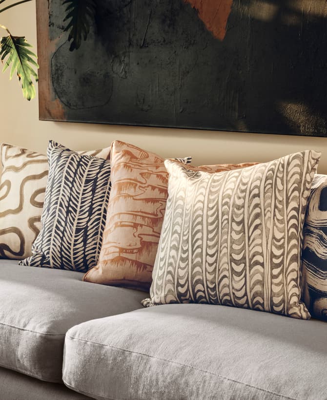 Five patterned cushions rest on a grey sofa, with a green potted plant and abstract wall art in the background.