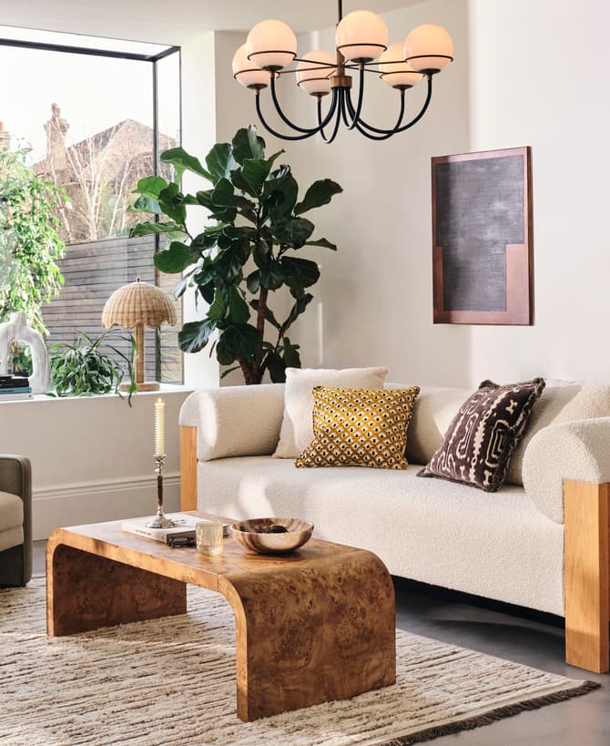 A cozy living room features a beige sofa and grey armchair with decorative pillows, centered around a wooden coffee table. Large windows, indoor plants, and art pieces create a welcoming ambiance.