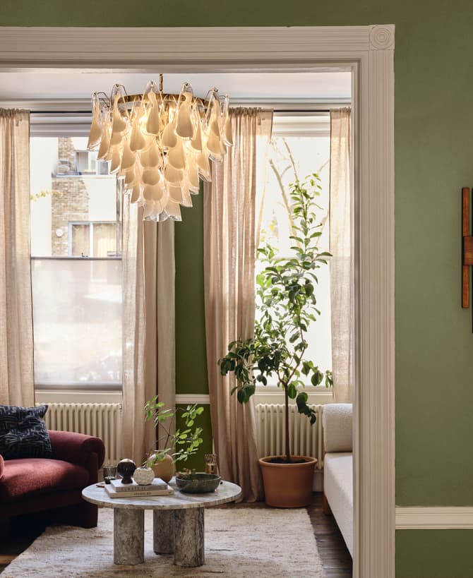 Chandelier hanging in a living room with a green armchair, coffee table, potted plants, and wall art. The space is softly lit with cream curtains and green walls.